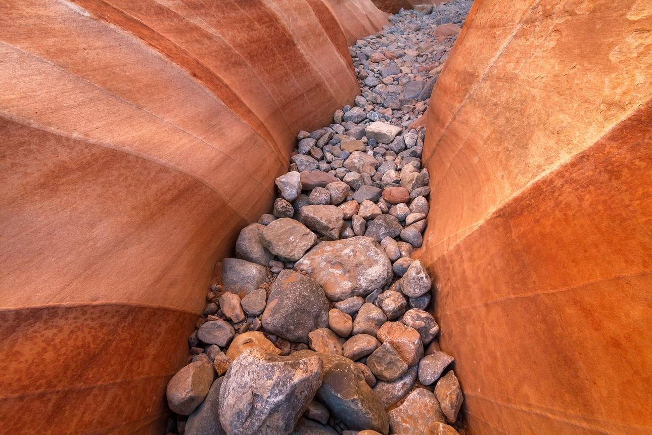 Slot canyon with smooth red rock walls and scattered stones along a narrow desert passage.