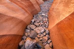 Slot canyon with smooth red rock walls and scattered stones along a narrow desert passage.